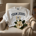 TEAM JESUS Collegiate Sweatshirt draped over linen armchair with magnolia branch
